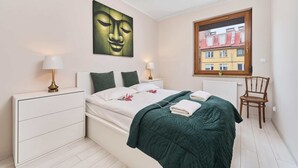 Une chambre à coucher comprenant un lit, des tables de chevet, une fenêtre donnant sur des immeubles et une œuvre d’art accrochée au mur.