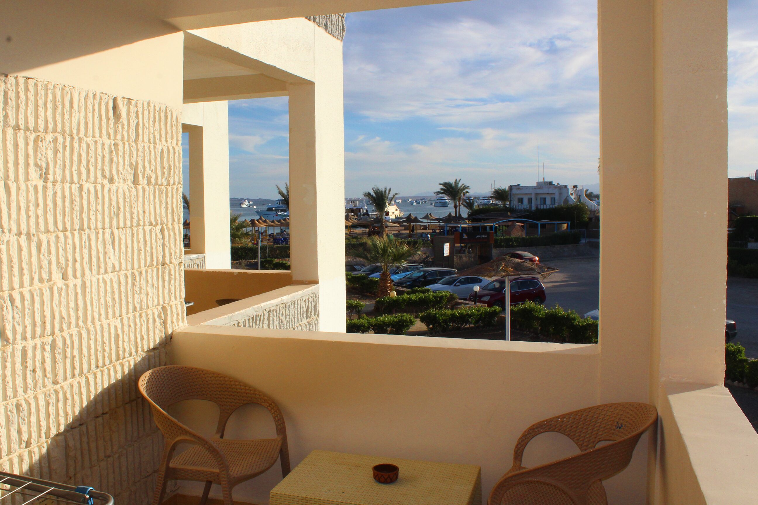 A balcony with wicker chairs and a table, overlooking a marina with boats and parked cars.