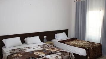 Habitación de hotel con dos camas, cabecera de madera, mesita de noche con lámpara y ventana con cortinas.