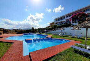 Una piscina con borde de ladrillo rojo, rodeada de sillones de descanso y un edificio con balcones.