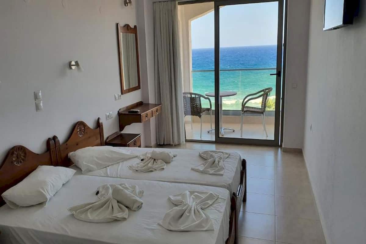 Een hotelkamer met twee bedden, een bureau, een stoel en een balkon met uitzicht op zee.