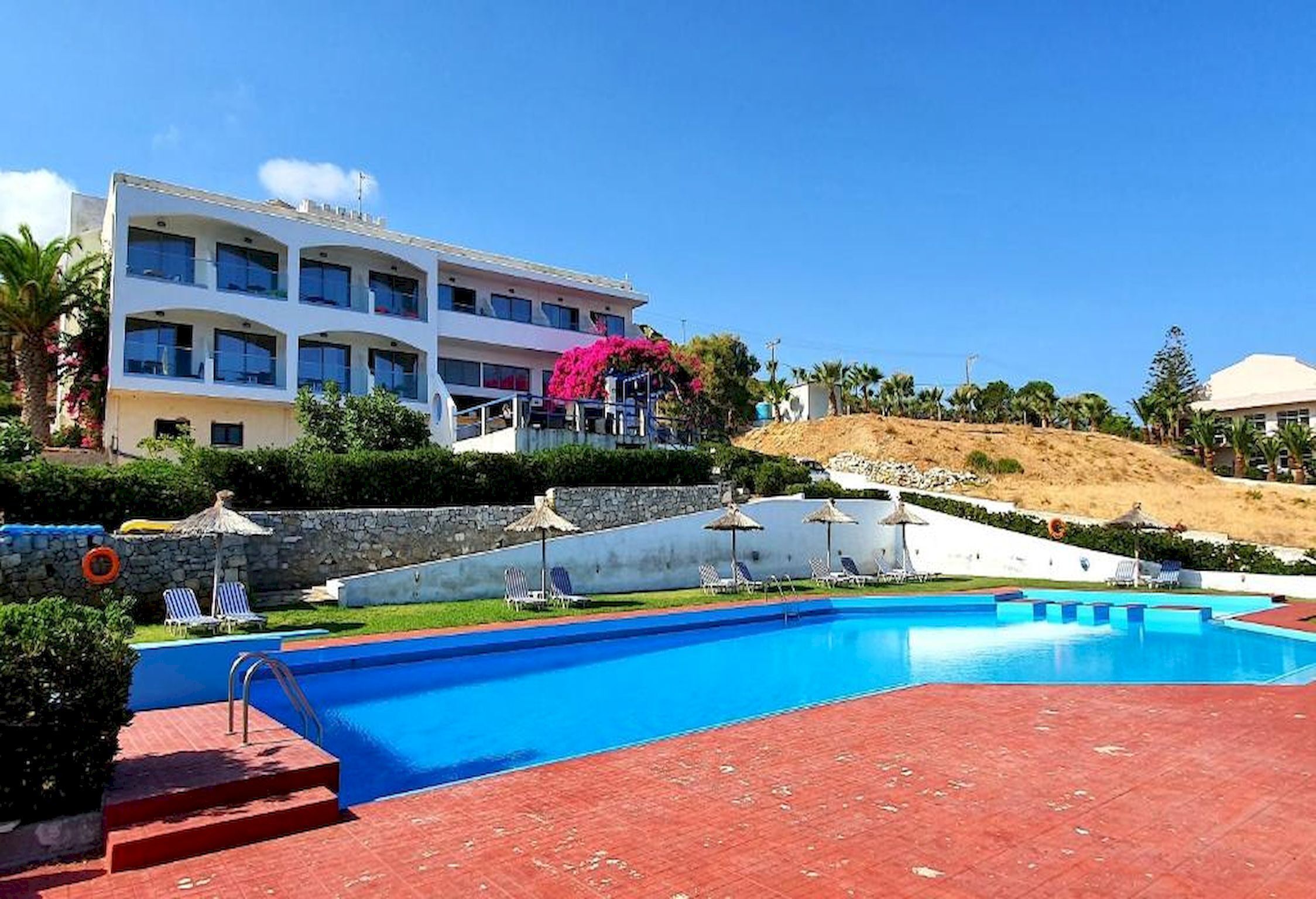 A swimming pool with a red-tiled deck, lounge chairs, and a white building with balconies.