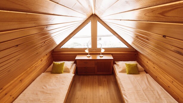 Un dormitorio acogedor en el ático con dos camas, paredes de madera, una mesita de noche pequeña y una ventana.