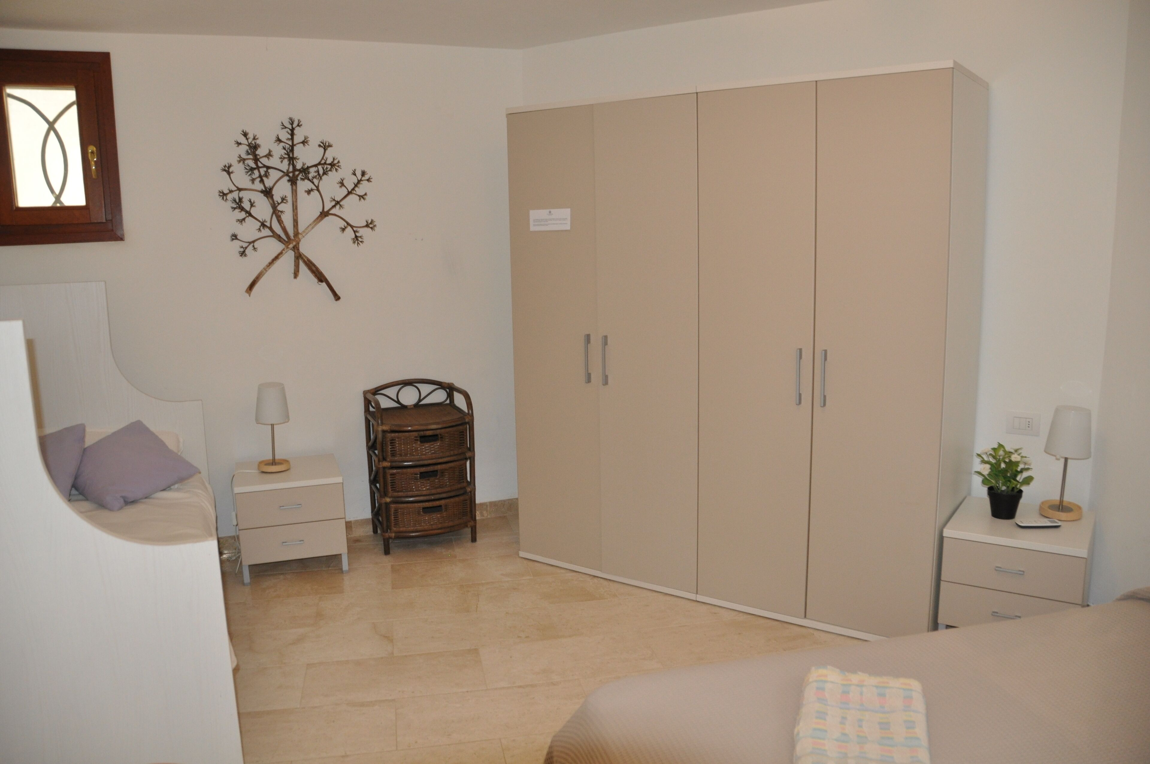 A bedroom with a bed, a wardrobe, a nightstand, a lamp, and a decorative tree.