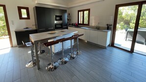 A modern kitchen with a central island, dark cabinetry, and a large window providing a view of the outdoors. - Villa Carla (Olmeto)