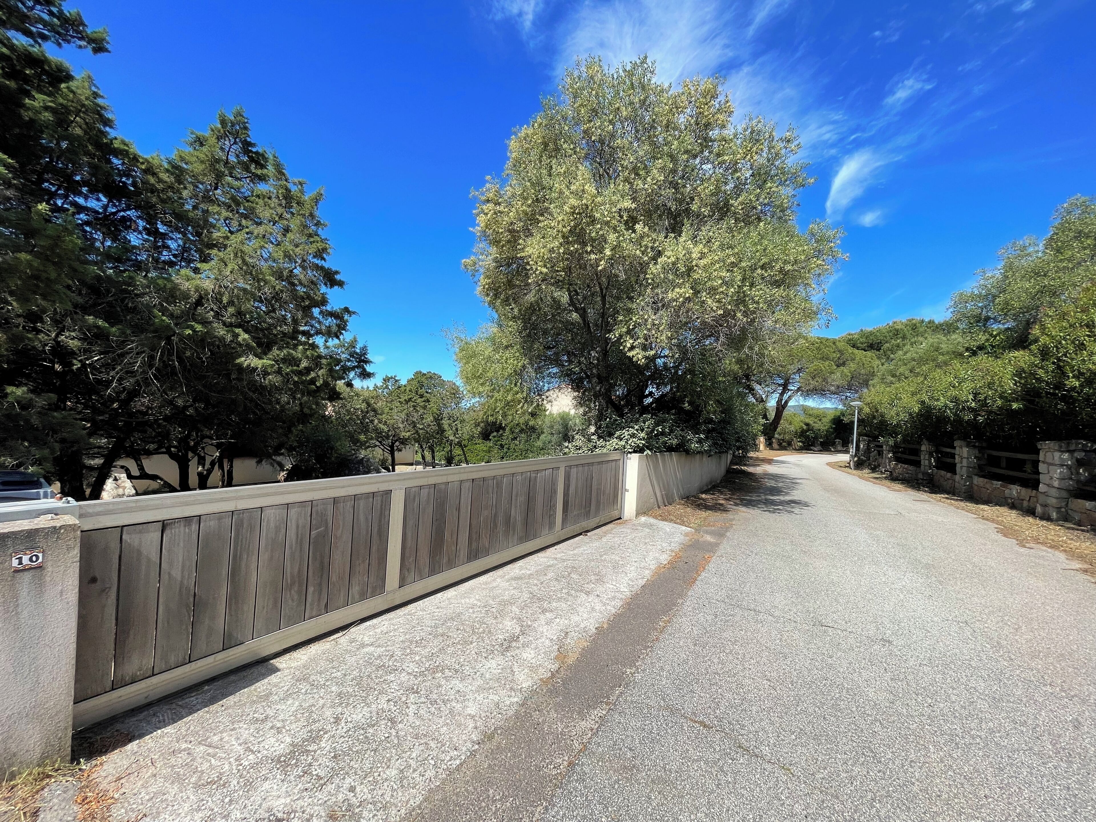 A paved road flanked by a wooden fence and stone wall, leading to a residential area with trees and a clear blue sky.