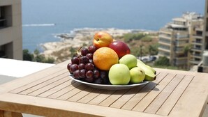 A plate of fruit on a wooden table with a view of the sea and buildings in the background. - Rawsheh 51 (Beirut)