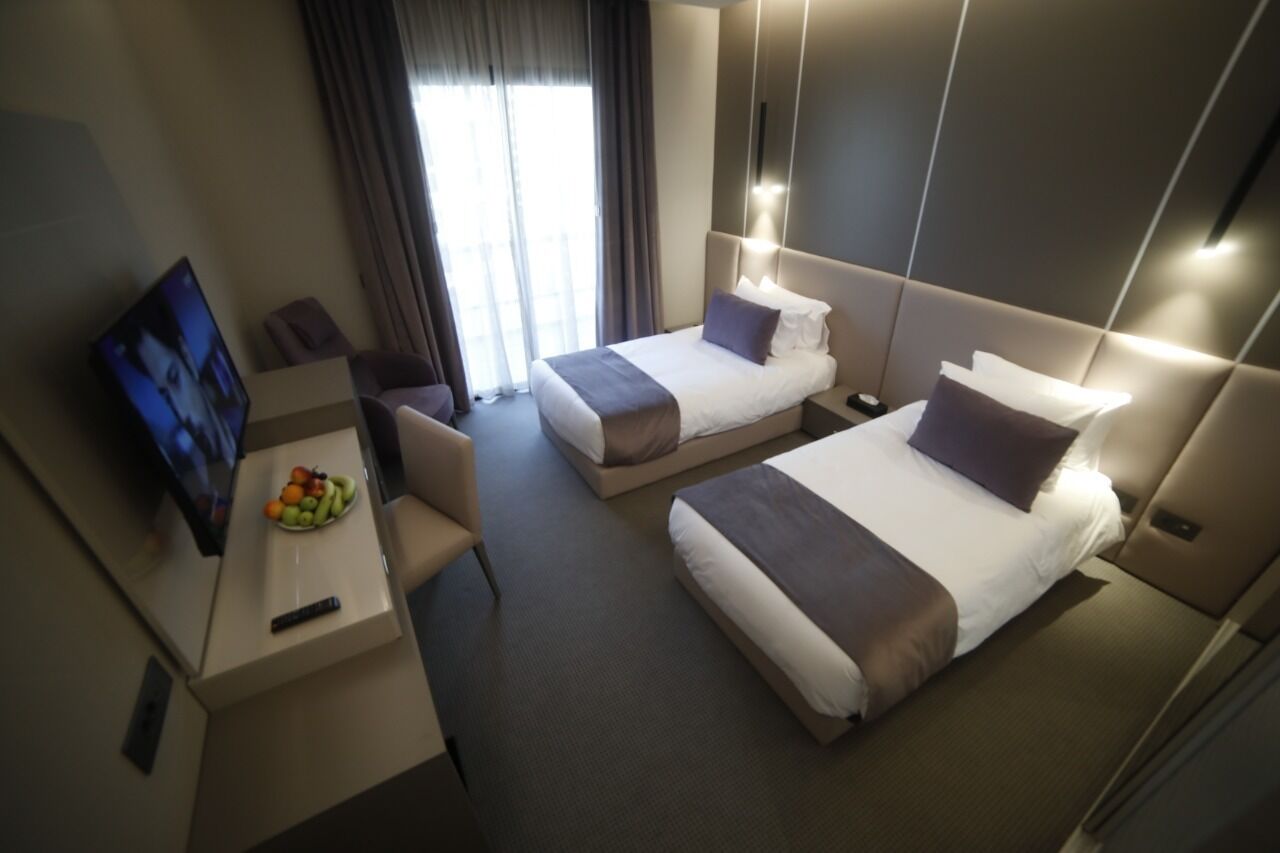 Habitación de hotel con dos camas, un escritorio con frutero, televisor y ventana con cortinas.