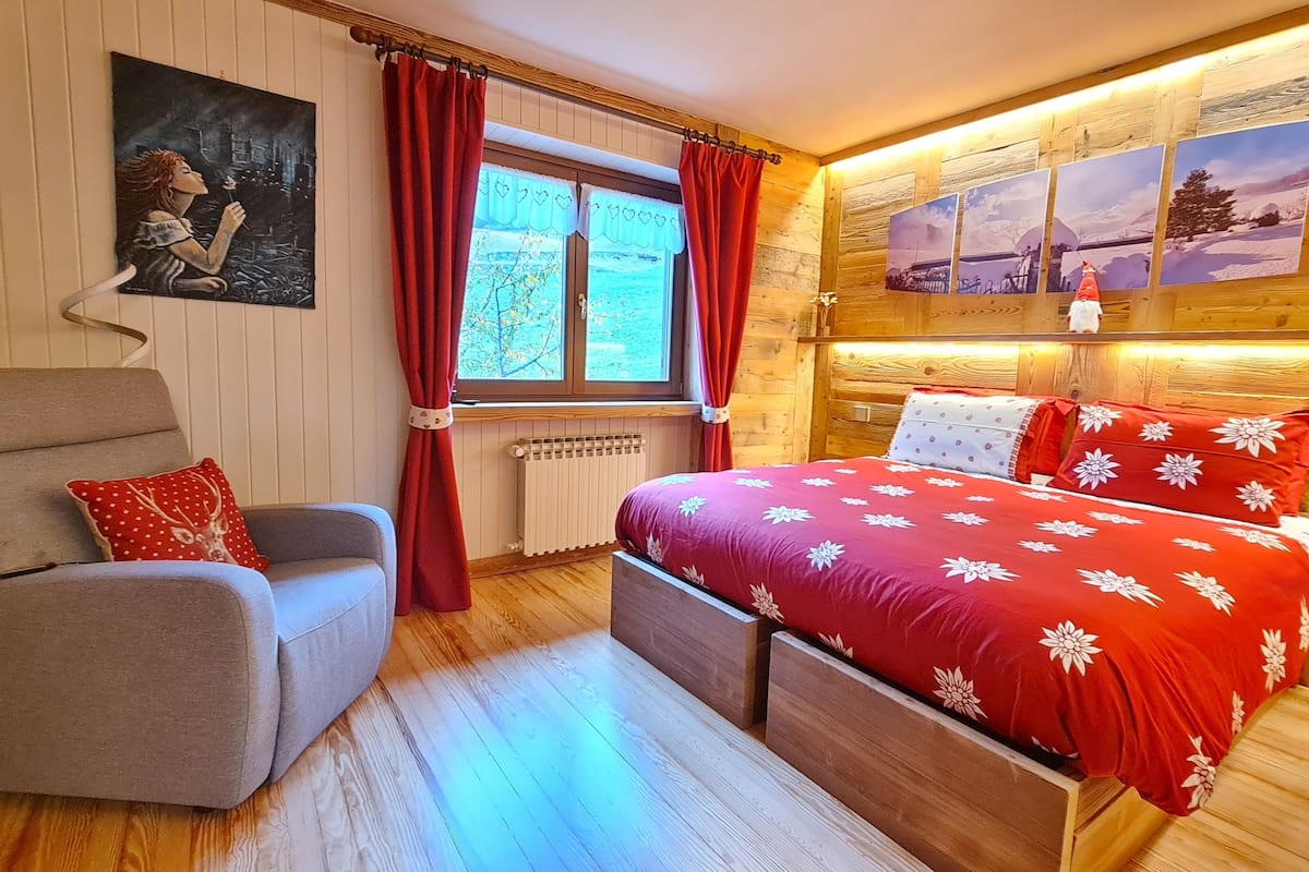 A cozy bedroom with a bed, a chair, a window with curtains, and a wall with framed pictures.