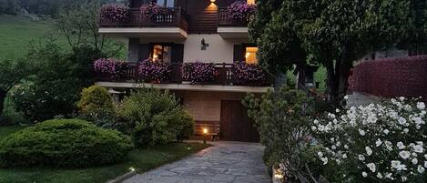 A two-story building with a balcony on each floor, surrounded by well-maintained gardens and a paved pathway leading to the entrance.