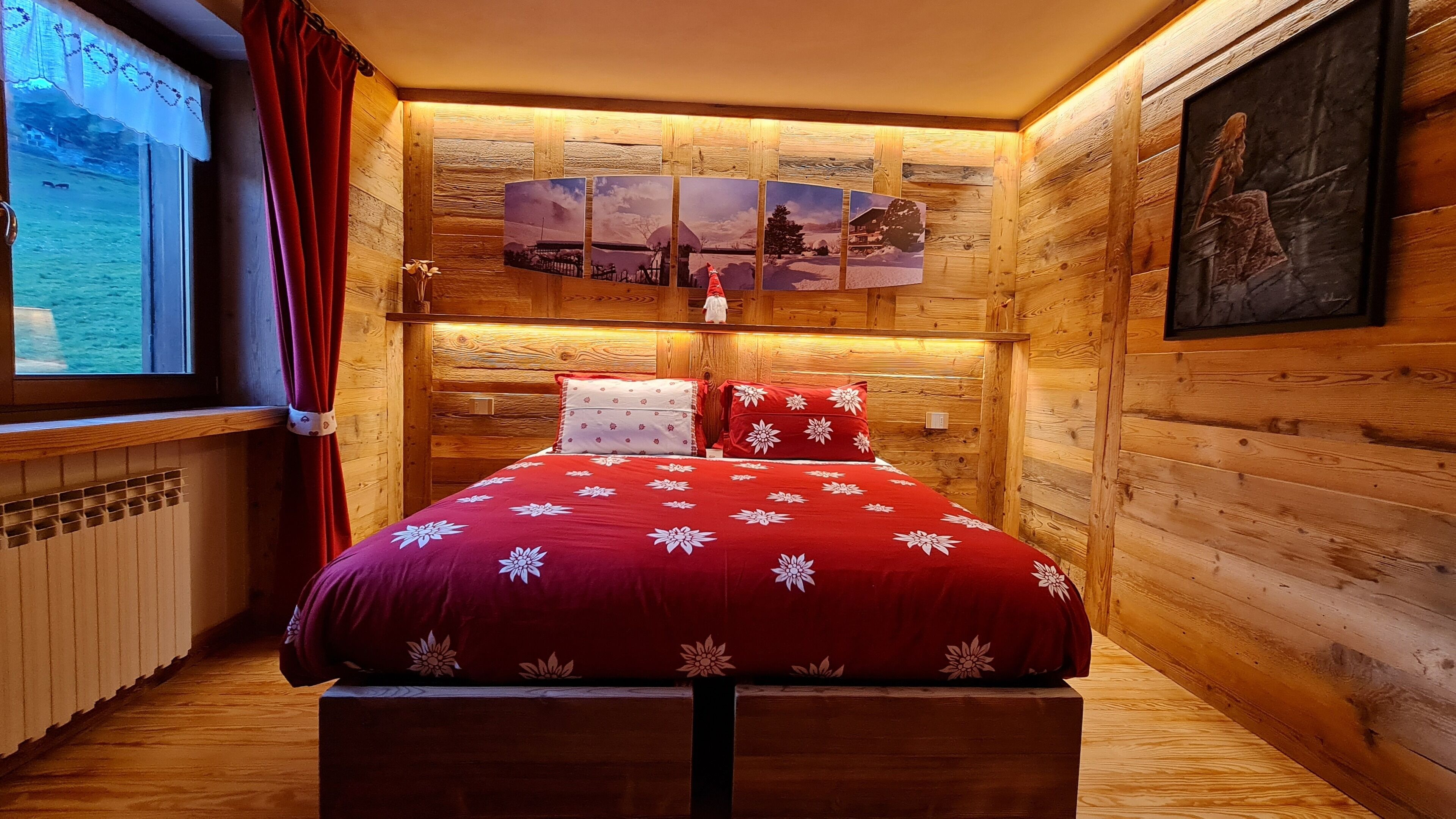 A cozy wooden room with a bed, a window with a view of a snowy landscape, and a series of framed pictures on the wall.