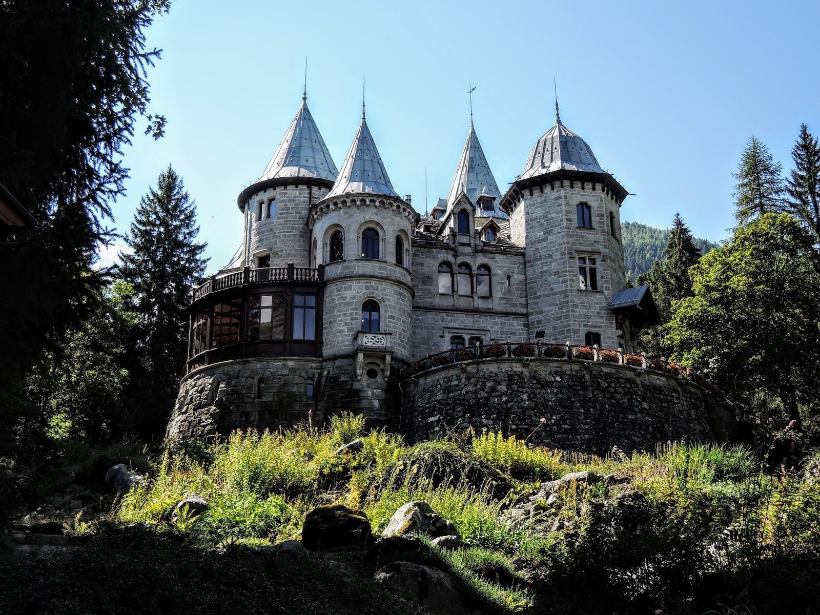 A castle-like building with multiple towers and a stone facade, surrounded by trees and rocky terrain.
