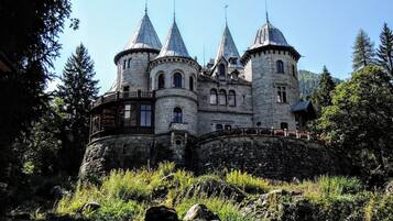 A castle-like building with multiple towers and a stone facade, surrounded by trees and rocky terrain.