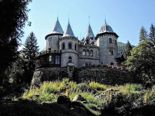 A castle-like building with multiple towers and a stone facade, surrounded by trees and rocky terrain.