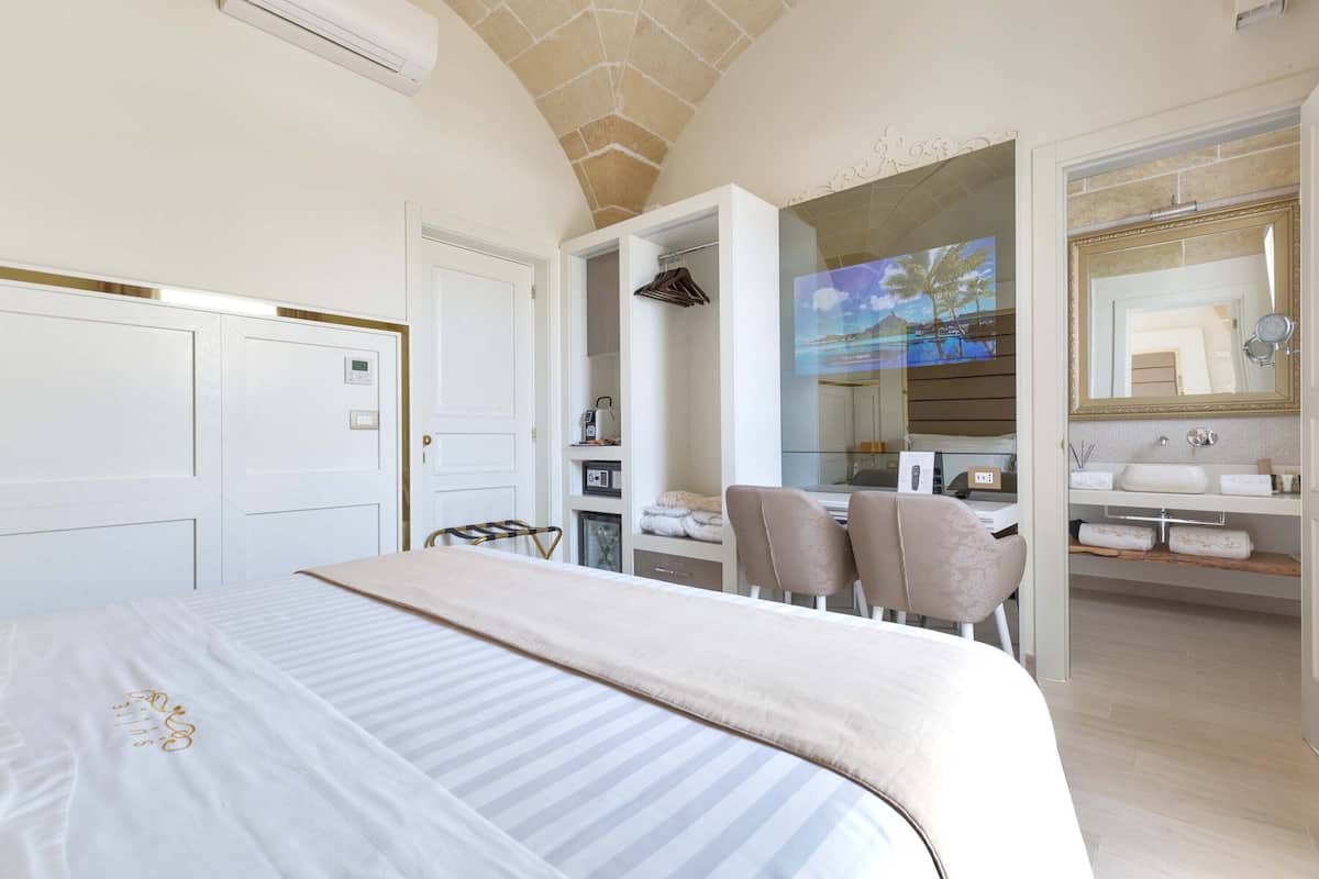 Una habitación de hotel moderna con una cama grande, un escritorio con sillas, un baño con espejo y lavabo, y un televisor que muestra una escena de playa.