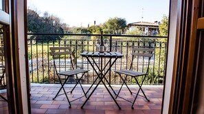 Un balcón con una mesa, dos sillas, una botella de vino y vistas a un jardín y edificios.