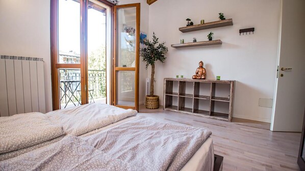 A bedroom with a bed, a potted plant, a wooden console table, and a window with a view of a balcony. - Appartamento Il Sole (Polpenazze del Garda)