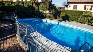 Une piscine entourée d’une rampe en métal, bordée d’un chemin pavé et d’une végétation luxuriante.