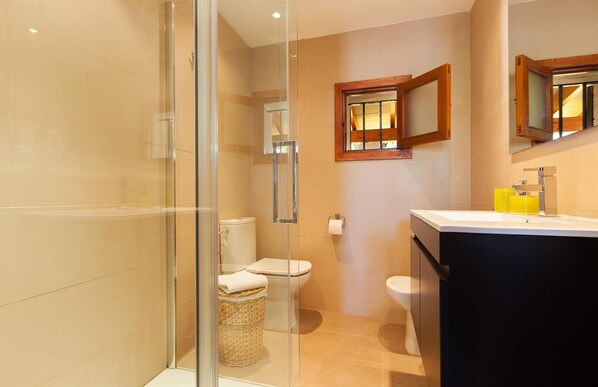 Un baño con cabina de ducha de vidrio, dos inodoros, un lavabo con espejo y una ventana con marco de madera.