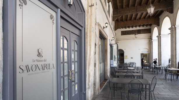An outdoor seating area with tables and chairs, a stone-paved floor, and a building with a sign reading "Le Stanze del Savonarola." - Le stanze del Savonarola  (Ferrara)