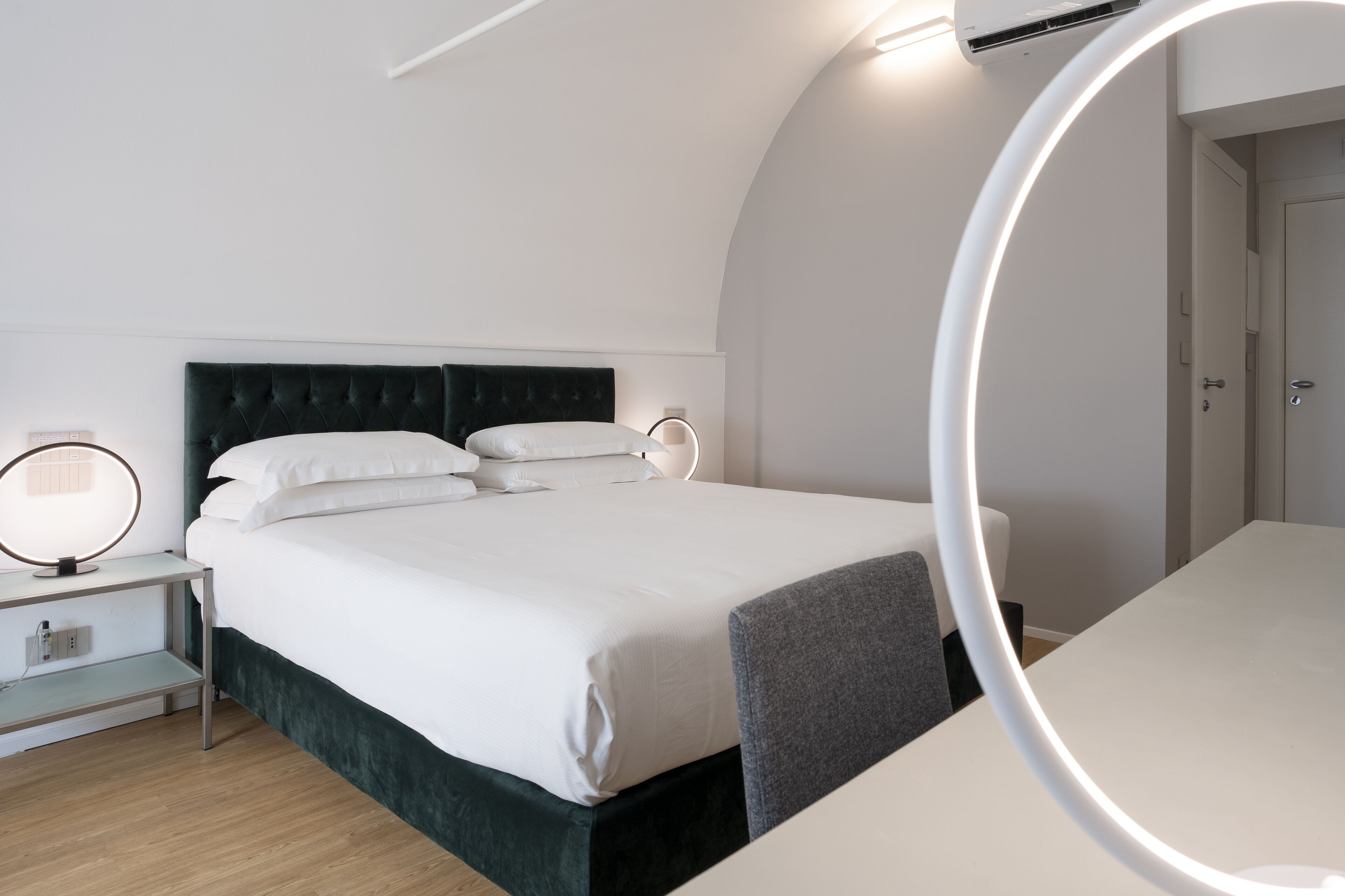 A modern hotel room with a neatly made bed, bedside table with a lamp, and a large mirror.