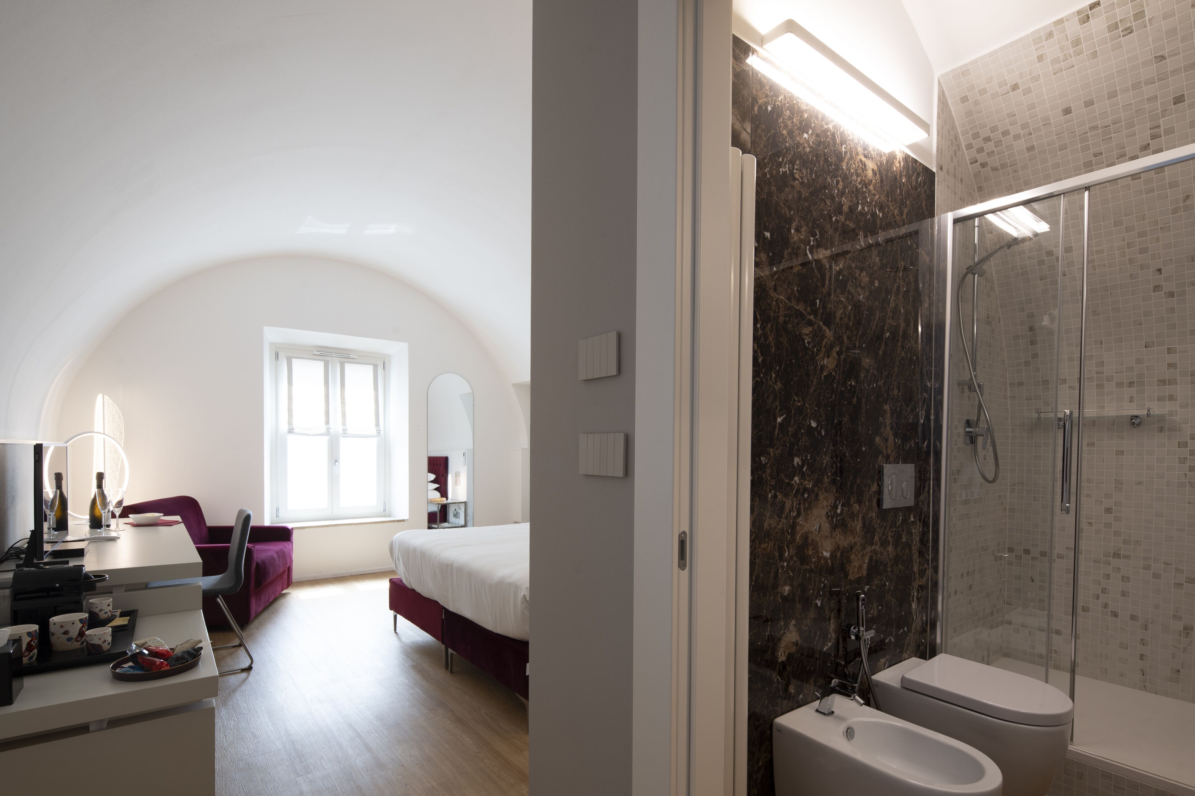 A modern bedroom with a large bed, a desk with a computer, and a chair. There is a bathroom with a shower and a toilet.