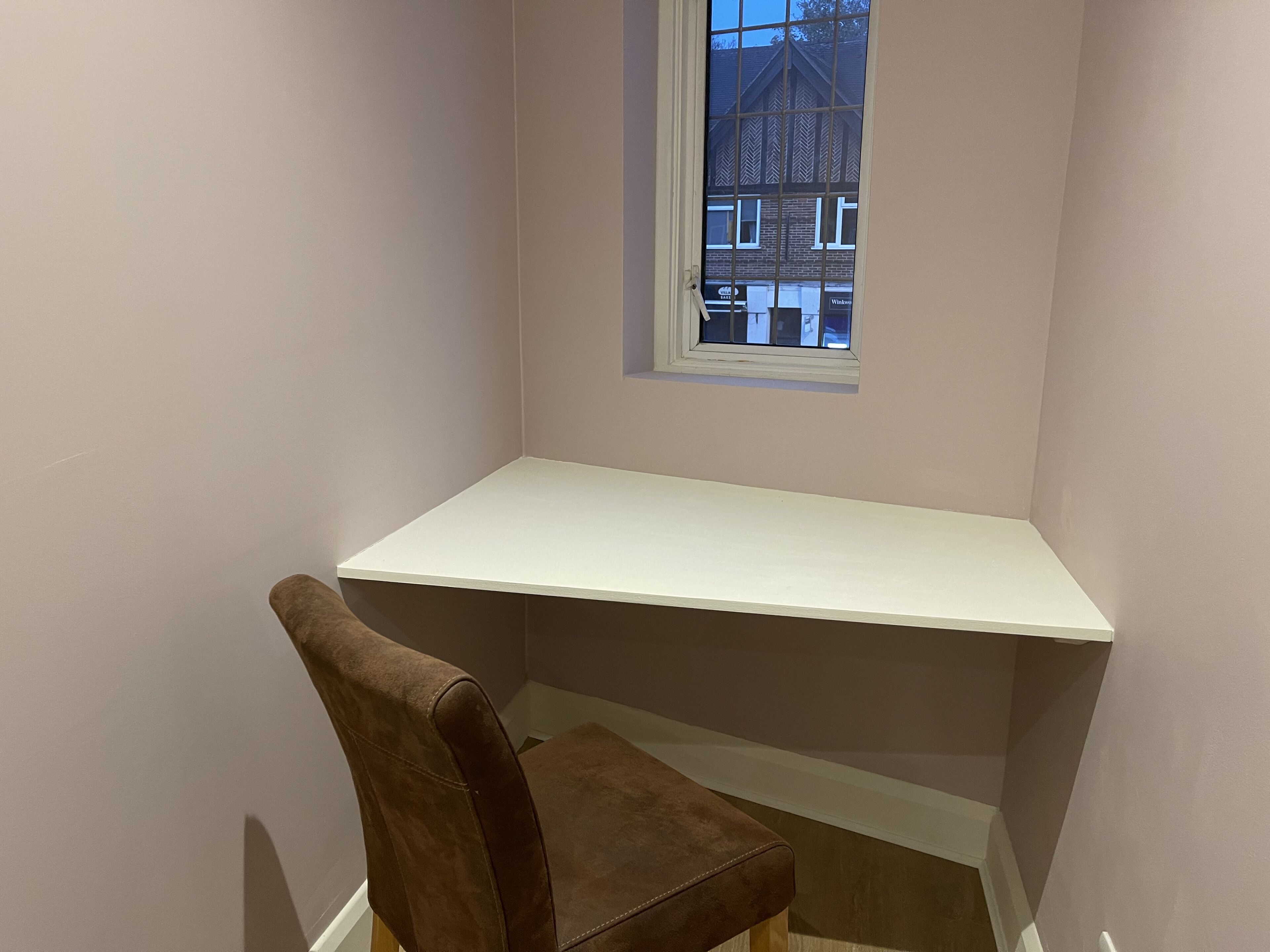 A corner room with a white desk, a brown chair, and a window overlooking a building.