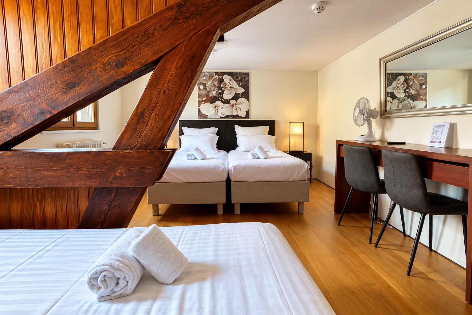 A hotel room with a bed, two bedside tables, a desk with a chair, a large mirror, and a painting on the wall.