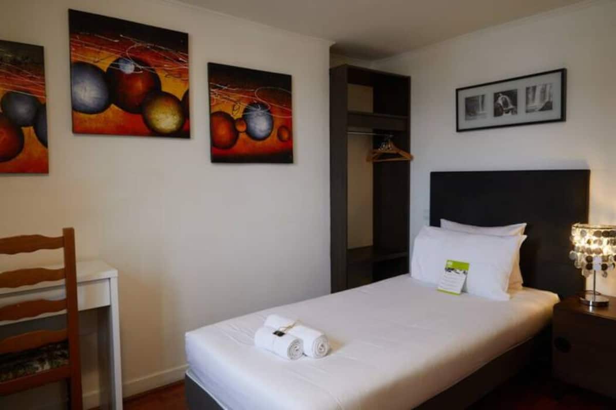 A hotel room with a bed, a chair, a wardrobe, and two paintings on the wall.