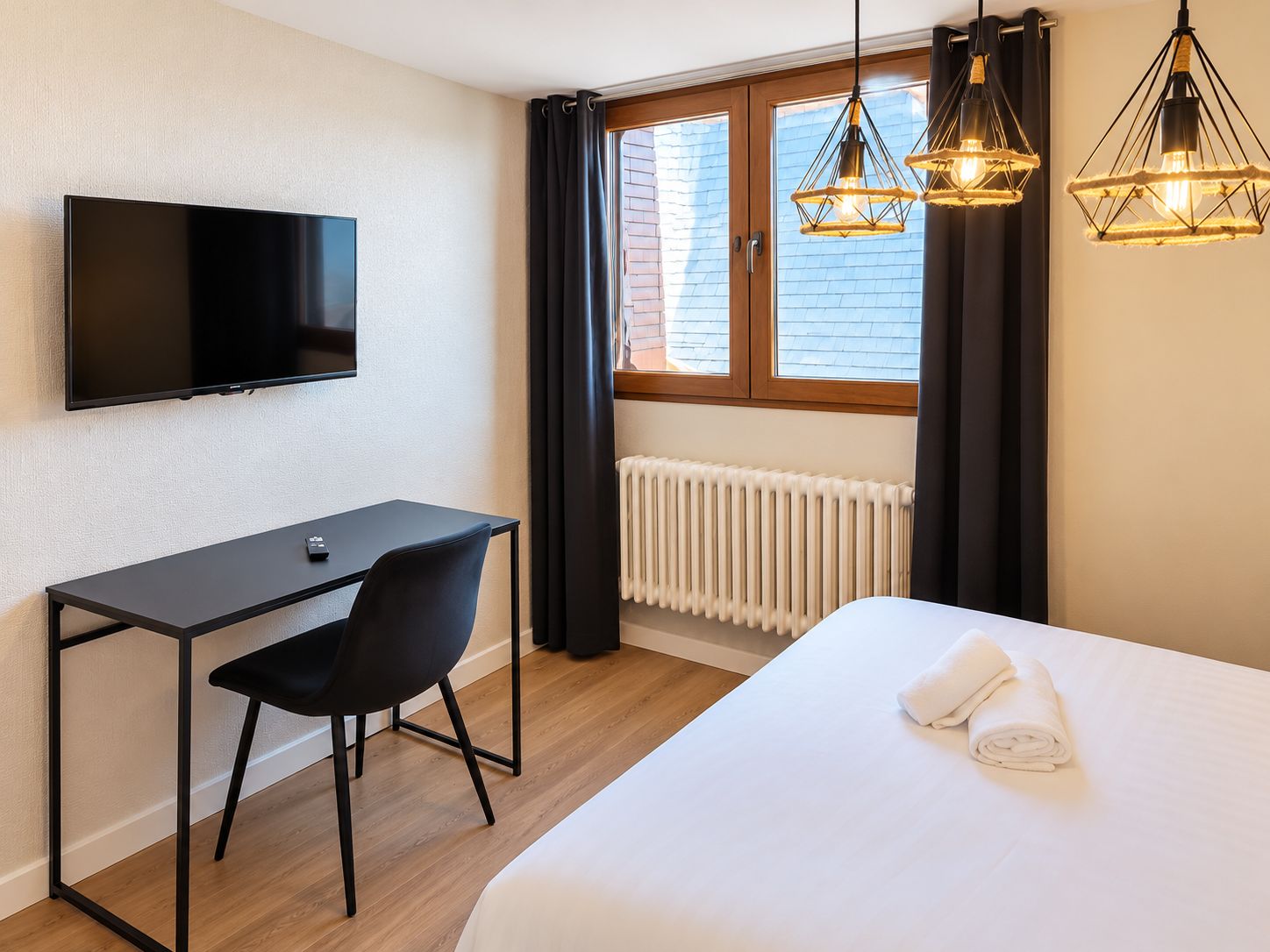 Una habitación de hotel con cama, escritorio con silla, televisor y dos lámparas colgantes.