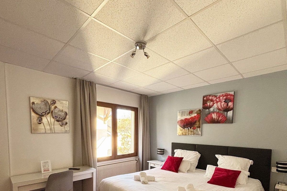 A hotel room with a large bed, a desk, and a chair. There is a window with curtains, a ceiling fan, and artwork on the walls.