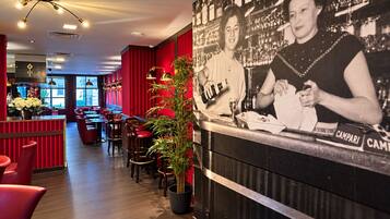 L’intérieur d’un restaurant présente des murs rouges, des tables et des chaises en bois. On y trouve une plante en pot et une photographie accrochée au mur représentant une personne.