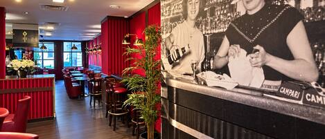 Een restaurantinterieur met rode wanden, houten tafels en stoelen. Er staat een potplant en er hangt een foto van een persoon aan de muur.