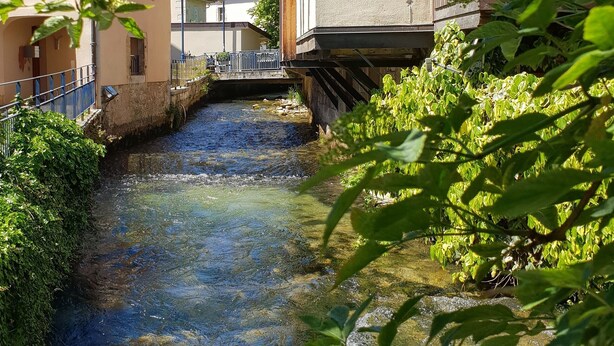 A narrow water channel with buildings on either side, a wooden bridge, and lush greenery. - Le Divona (Divonne-les-Bains)