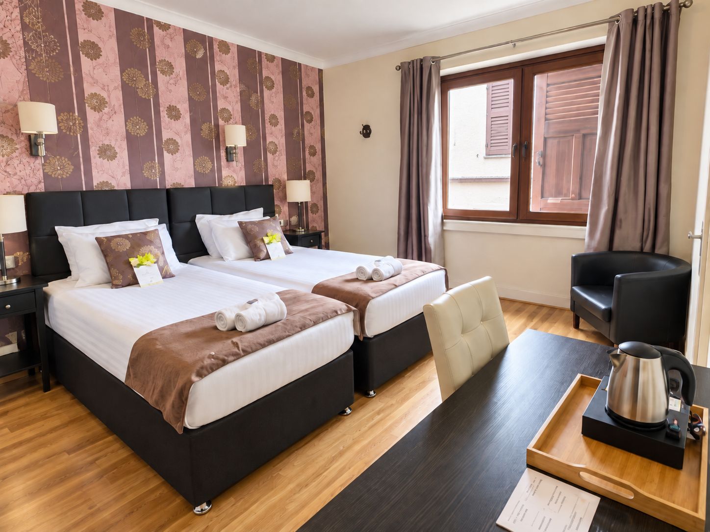 A hotel room with two beds, a desk with a kettle, a chair, and a window with curtains.