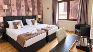A hotel room with two beds, a desk with a kettle, a chair, and a window with curtains.