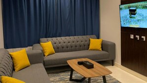 A modern living room with a grey sectional sofa, yellow cushions, a wooden coffee table, and a television mounted on a wooden panel. - Qimam Park Hotel 2 Abha (Abha)