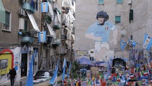 Une rue étroite, bordée de bâtiments des deux côtés, dont certains sont dotés de balcons et de climatiseurs. Sur l’un des bâtiments, on peut admirer une grande fresque représentant une personne en tenue de sport. Des drapeaux sont accrochés au-dessus de la rue, et une voiture est garée sur le bord.