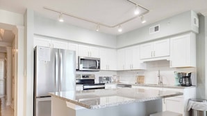 A modern kitchen with white cabinetry, a stainless steel refrigerator, a microwave, a stove, and a coffee maker. There are two white stools at the island.