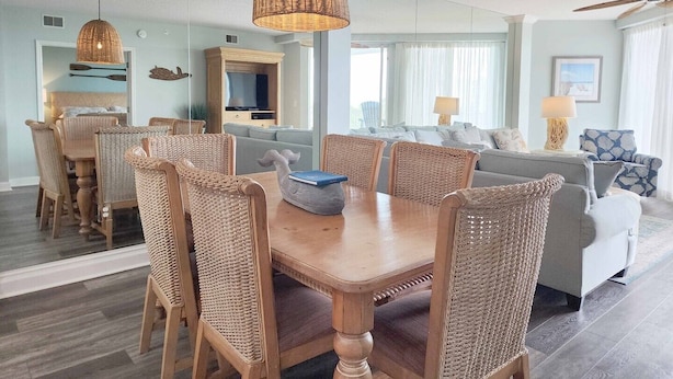 A dining area with a wooden table and wicker chairs, a living room with a sofa and a TV, and a ceiling fan.