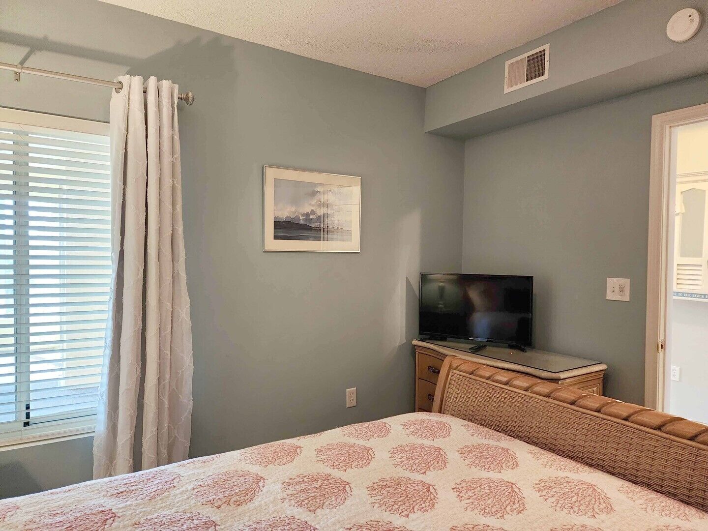 A bedroom with a bed, a television on a wooden dresser, a framed picture on the wall, and a window with curtains.