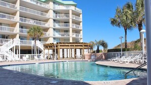 A pool area with a wooden deck, lounge chairs, and a building with balconies in the background.