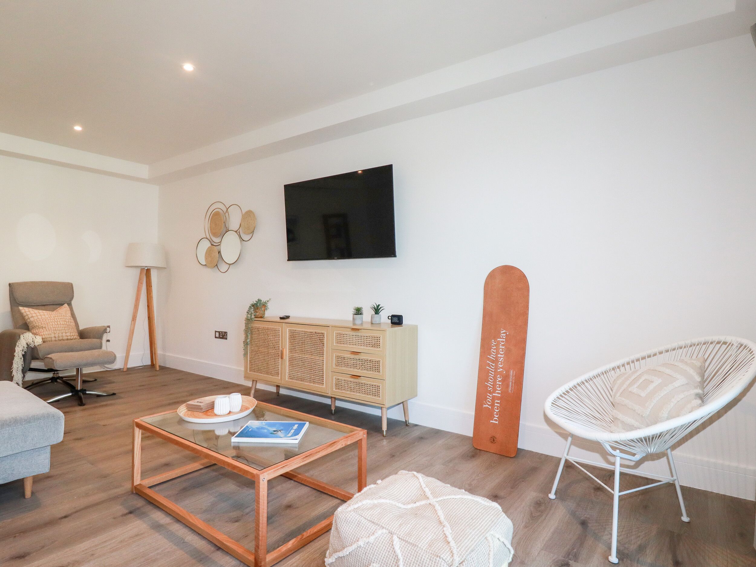 Ein modernes Wohnzimmer mit einer Couch, einem Sessel, einem Glastisch, einer Holzkommode und einem an der Wand befestigten Fernseher.