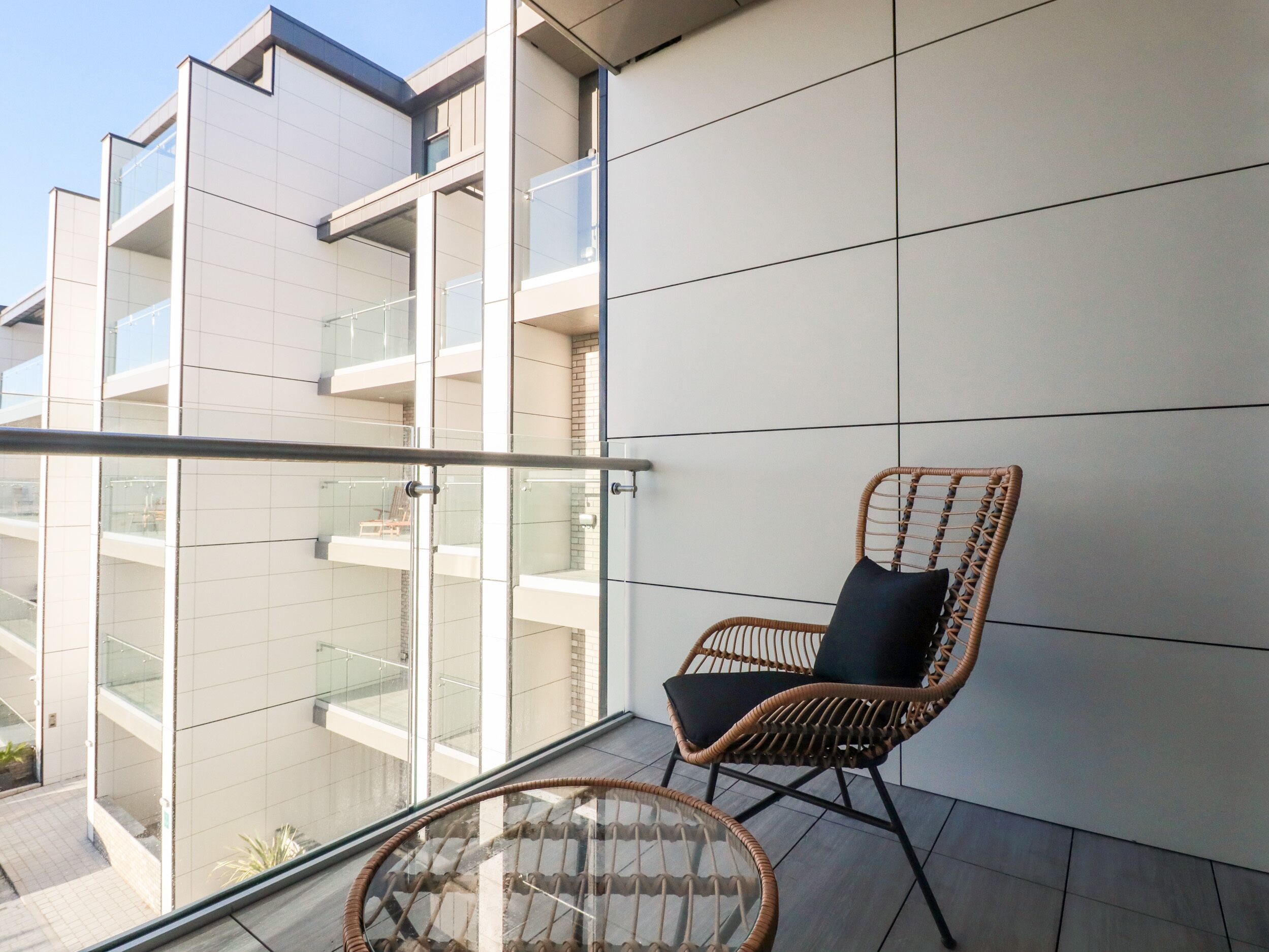 A balcony with a wicker chair and a glass table, overlooking a modern building with multiple balconies.