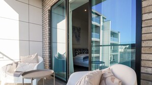 Un balcón con una silla blanca, una mesa pequeña y un sofá con cojines. Una puerta de cristal da a un dormitorio con cama y balcón.