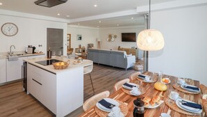 Una cocina moderna con una isla central, un comedor con mesa de madera dispuesta para el desayuno y una sala de estar con sofá y televisor.