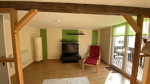 A room with wooden beams, a red chair, a black entertainment unit, and a large window with a view of a balcony. - Cozy vacation home at the foot of the Hoher Meißner (Meißner)