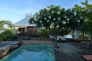 A pool area with a wooden deck, a small tree with white flowers, and a building with a sloped roof. - Le Farhé - Villa Harmonie Ilot Fortier (Petite Riviere Noire)