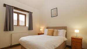A bedroom with a large bed, two bedside tables with lamps, a window with curtains, a radiator, and a framed picture on the wall. - Cinder Cottage (Worcester)
