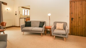 A cozy living room with a grey sofa, a smaller armchair, a wooden side table, and a floor lamp. A wooden door and a kitchen with a window are visible in the background. - Cinder Cottage (Worcester)